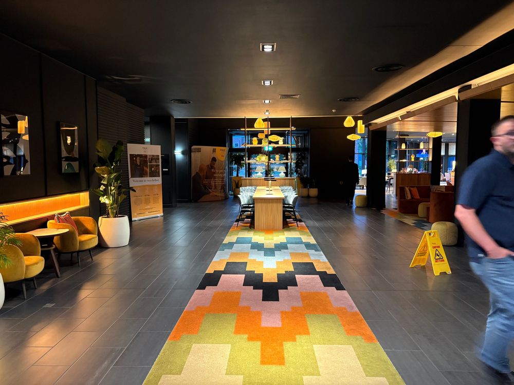 Modern hotel lobby with colorful geometric carpet runner, dark floors, and yellow accent furniture creating a welcoming space.