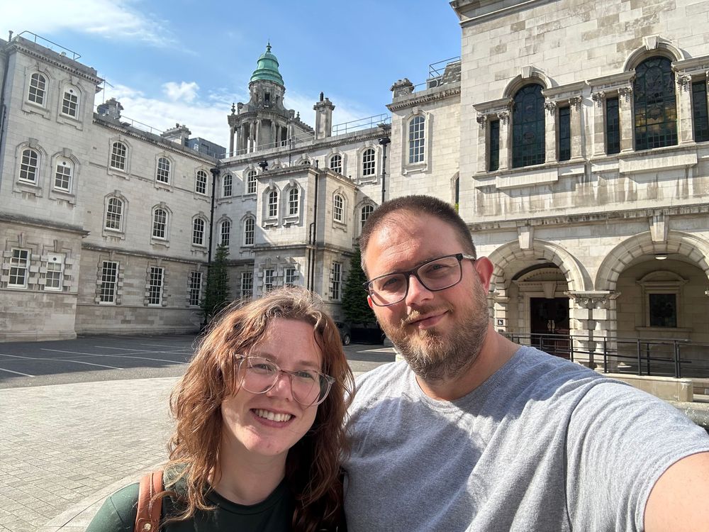 Two people take a selfie in front of an ornate stone building with a domed tower and classical architecture.