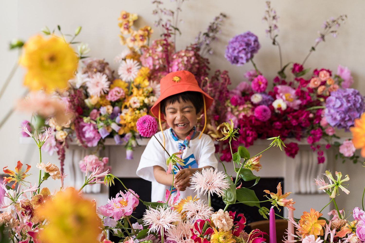 A young child wearing a pink hard hat sits among vibrant, colorful floral arrangements with a digital tablet.