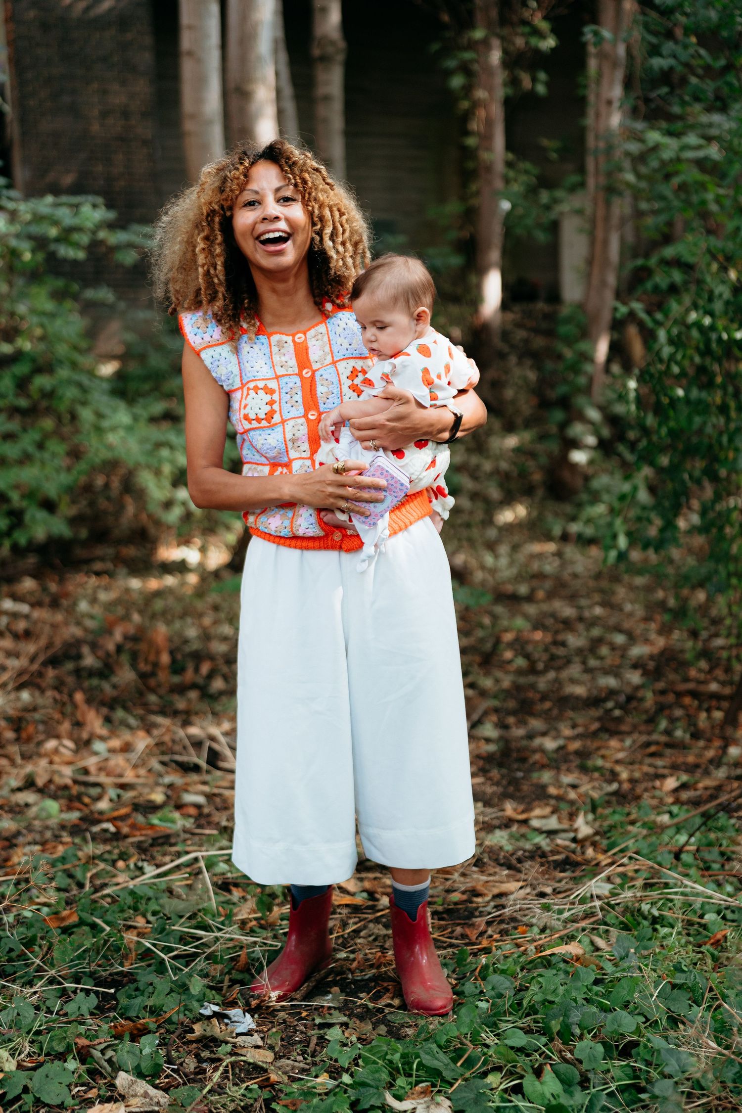 Happy mother and baby wearing coordinating orange and white outfits stand together in a wooded outdoor setting.