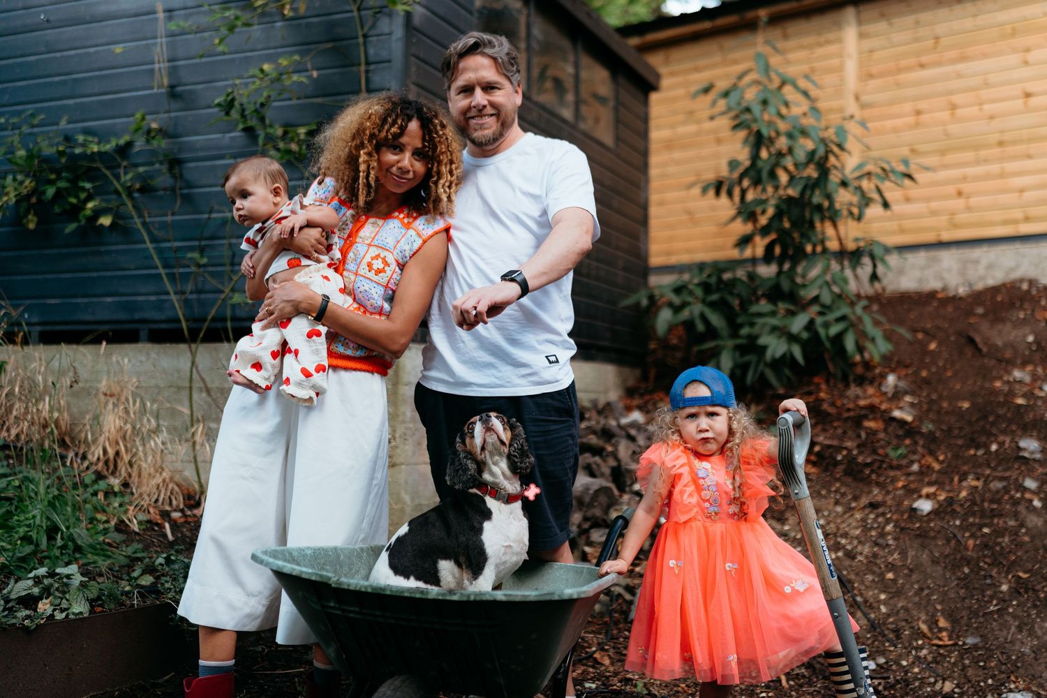 Family enjoys candid moment in garden with their small dog while casually posed around rustic wheelbarrow.