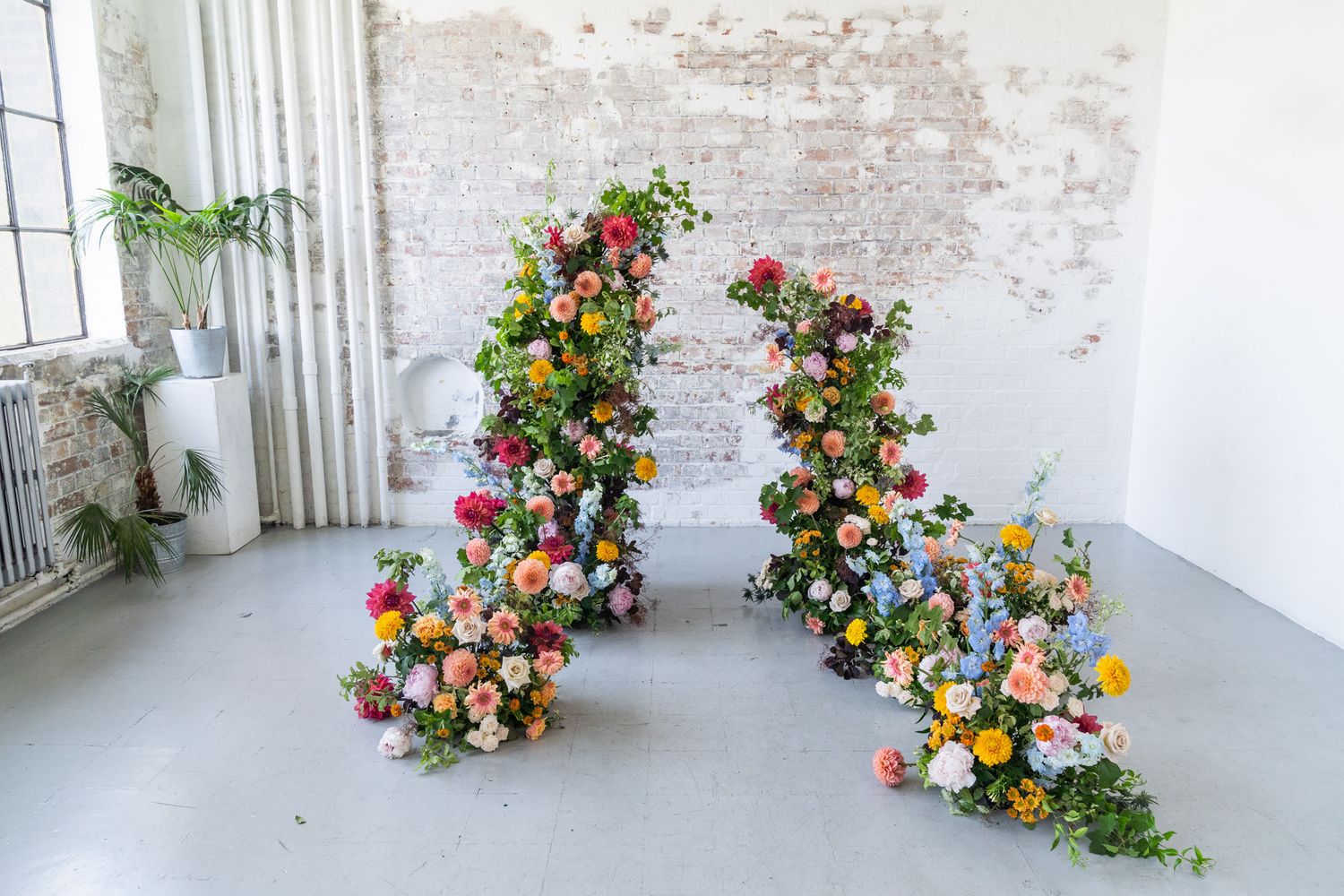 Two spiral floral arrangements with colorful blooms climbing upward against a white brick wall in a studio space.