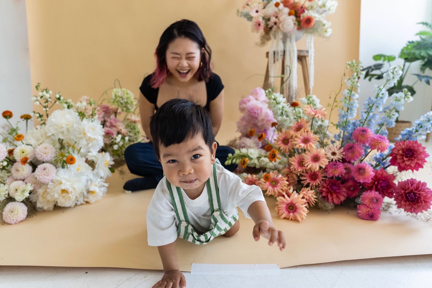 Two people pose playfully amongst a stunning array of colorful flowers against a beige backdrop during a photo session.