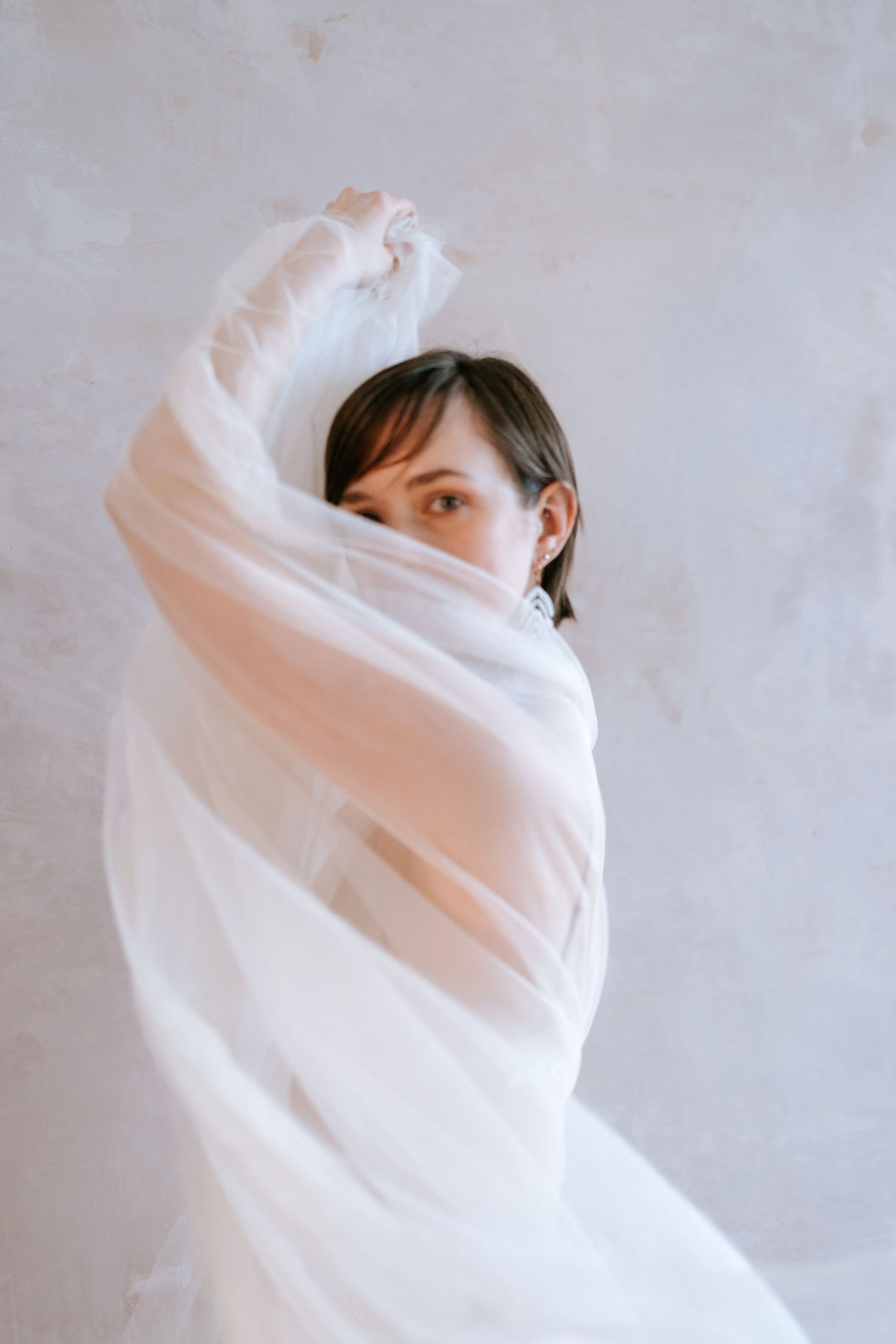 A person wrapped in flowing white fabric poses against a light background, creating an ethereal and dreamy effect.