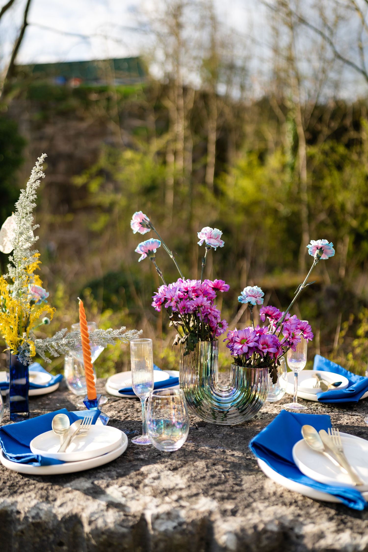 Romantic outdoor table setting with blue napkins and vibrant purple flowers in glass vases on a rustic stone surface.