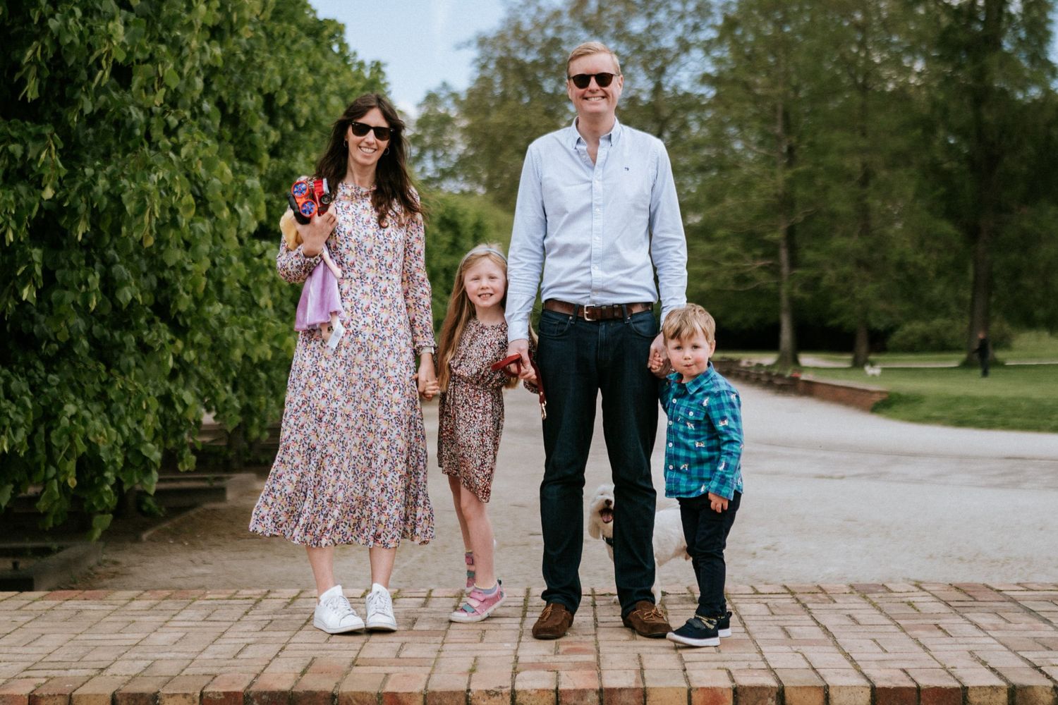 A family of four stands together on a brick pathway in a lush park setting during a sunny day.