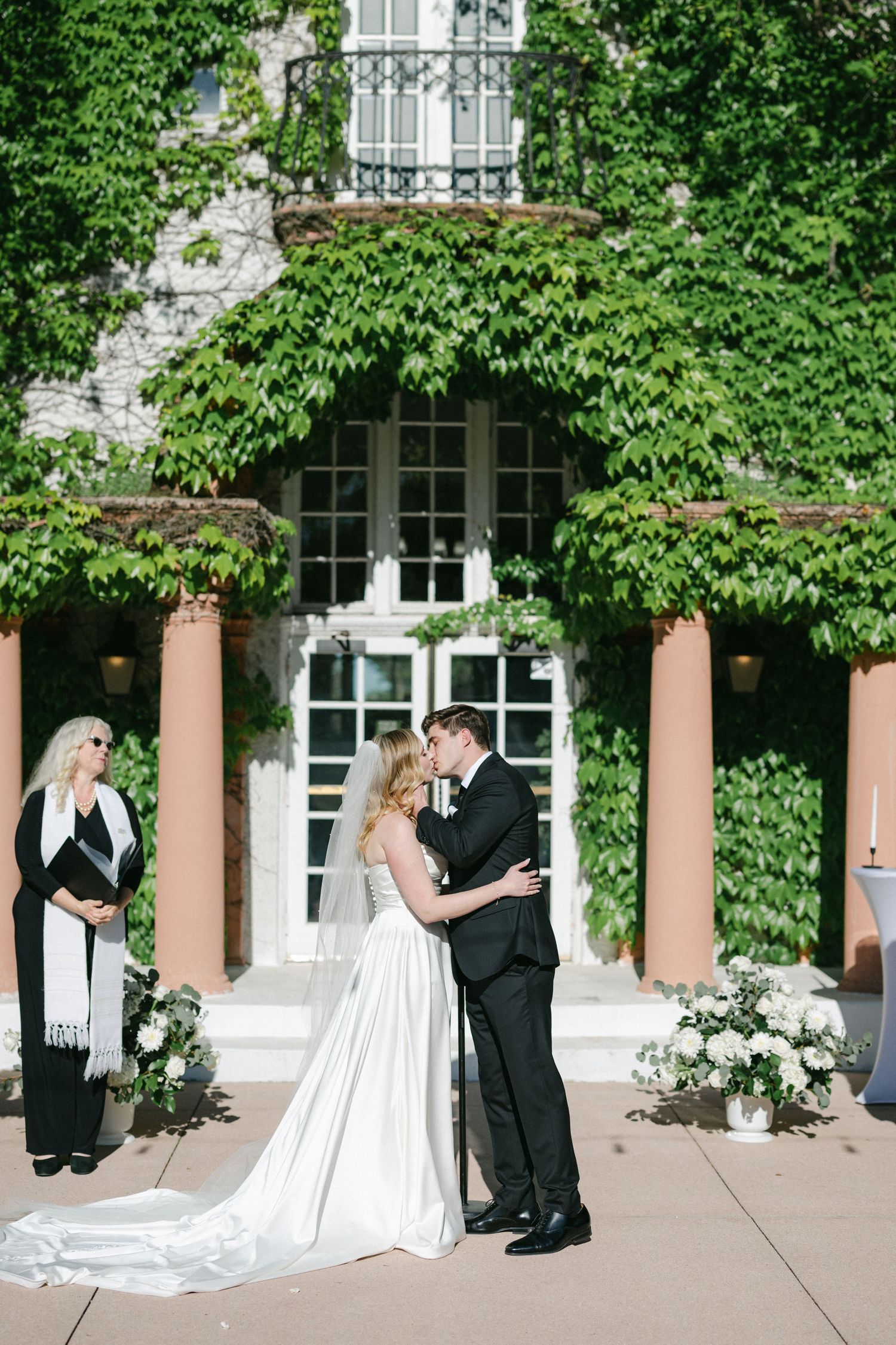 Wedding couple shares a romantic moment under an ivy-covered archway with pink columns at an elegant venue entrance.