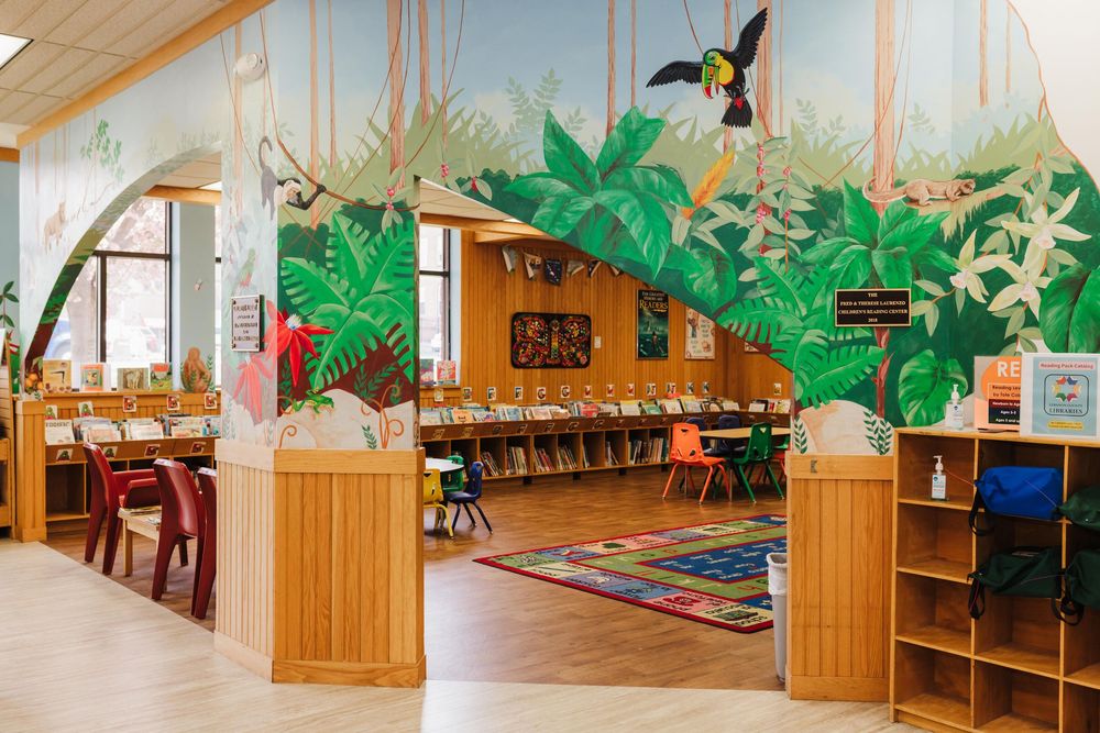 Kids library space decorated with tropical green paper palm leaves and wooden furniture features a colorful reading rug and bookshelves.