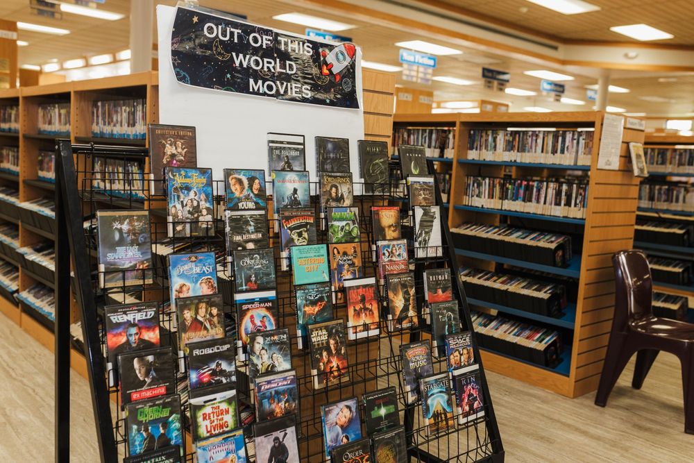 A display rack of DVDs and movies in a library featuring various titles under an 'Out of This World Movies' sign.