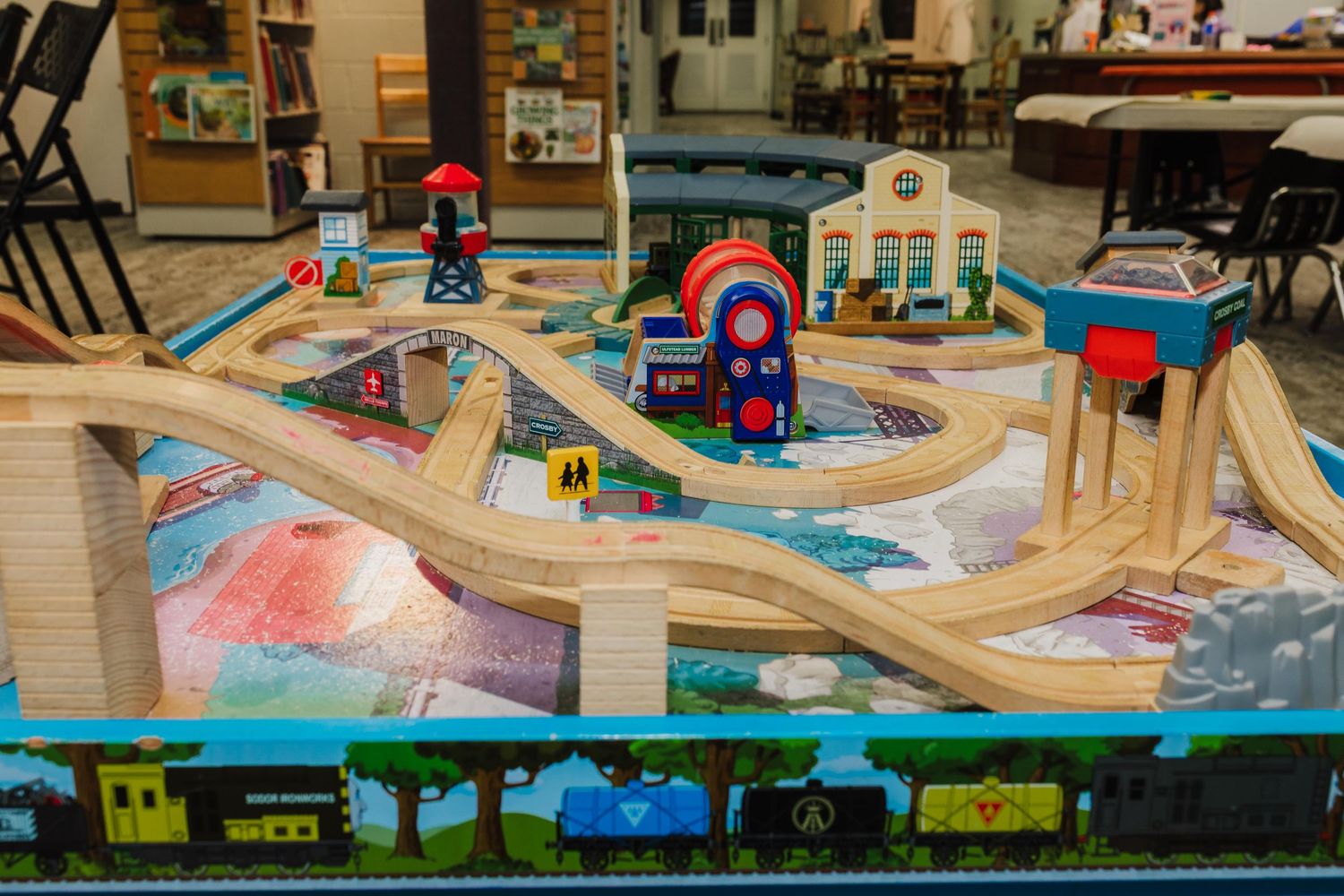Wooden toy train track setup with bridges and tunnels displayed on a play table with decorative train artwork below.