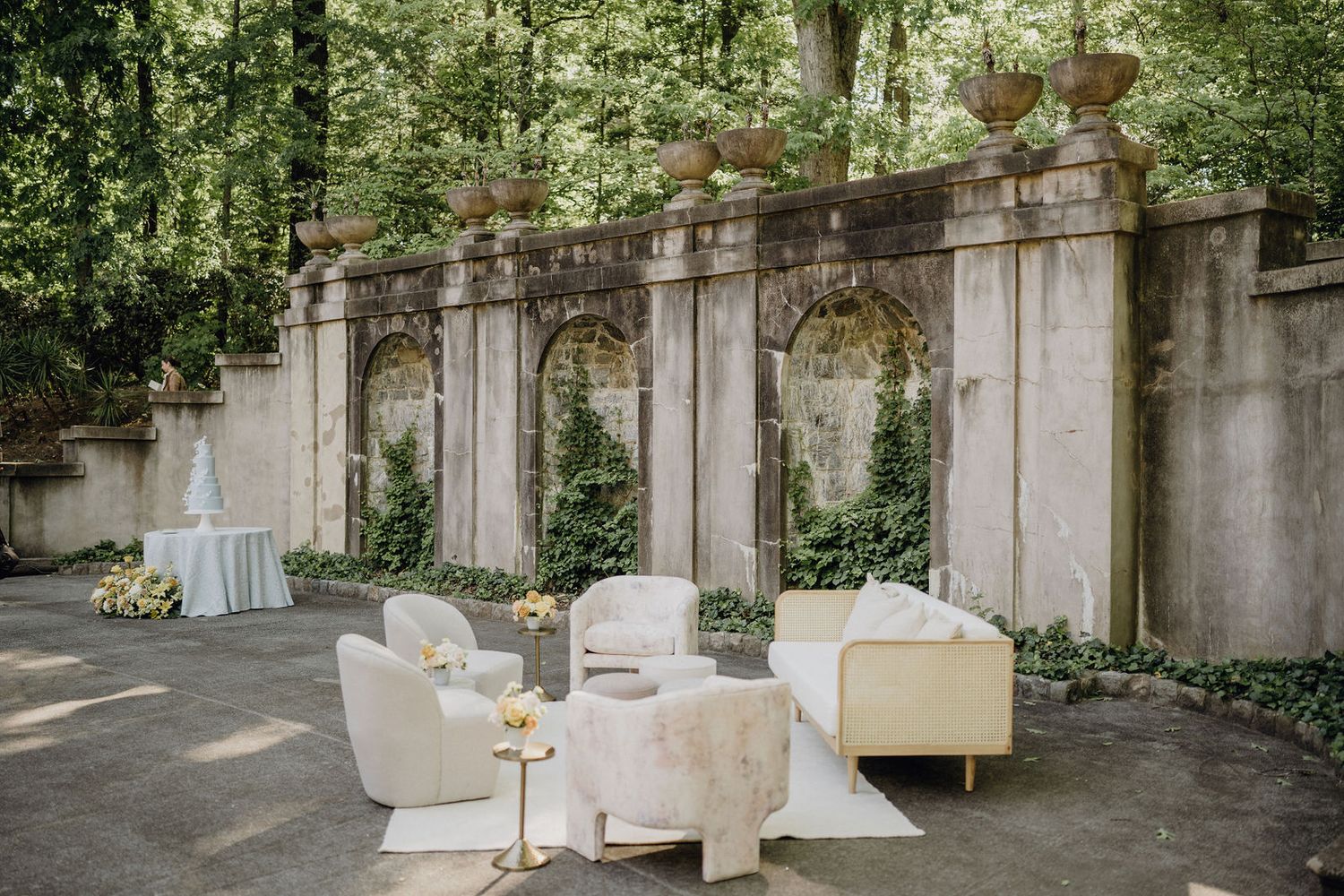 Elegant outdoor wedding lounge setup with white furniture against stone wall with climbing vines.