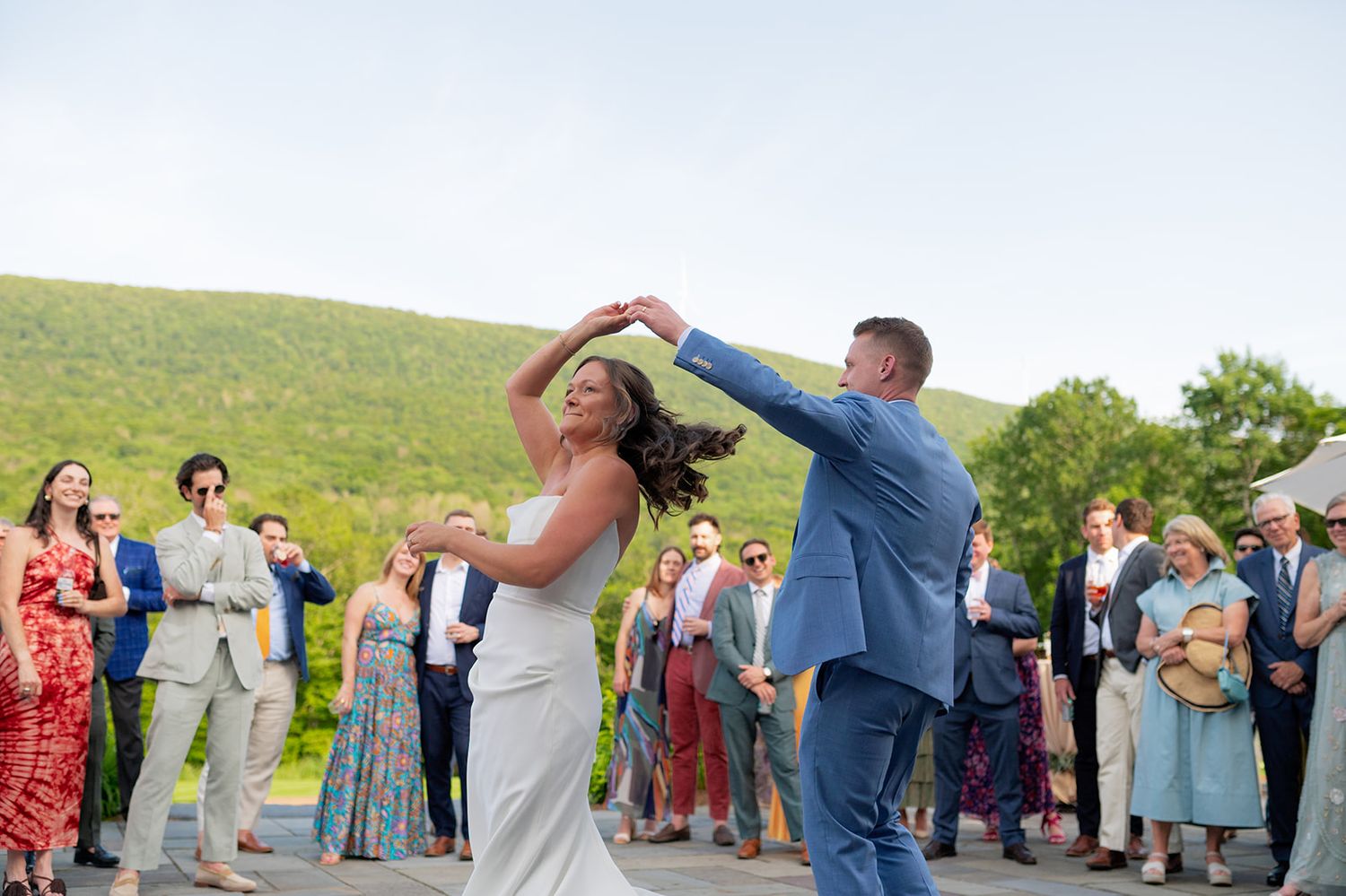 Wedding party dances in a conga line on a grassy hillside during an outdoor celebration.