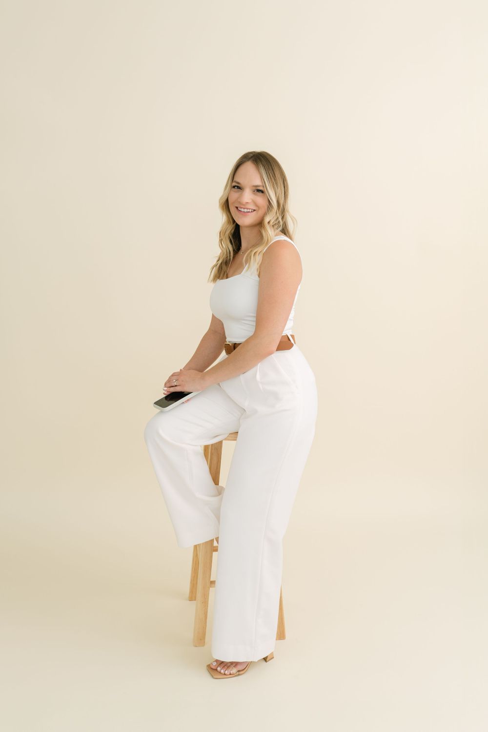 Woman in casual white outfit sitting on wooden stool against beige backdrop in studio setting.