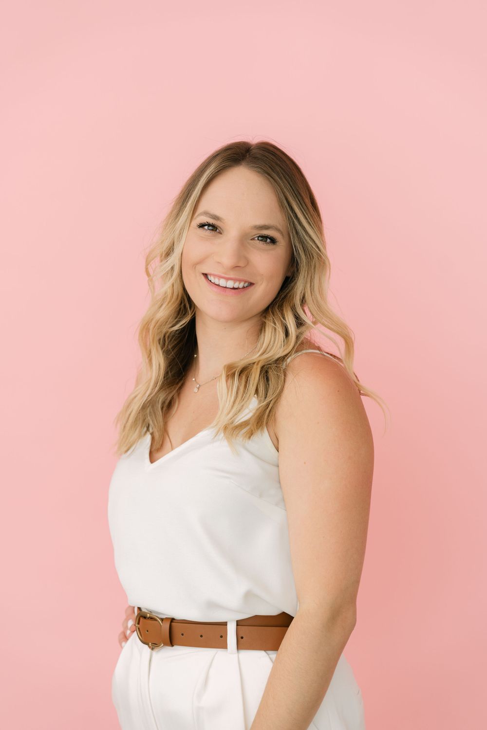 Professional portrait against pink background features a person in white outfit with brown belt smiling warmly.
