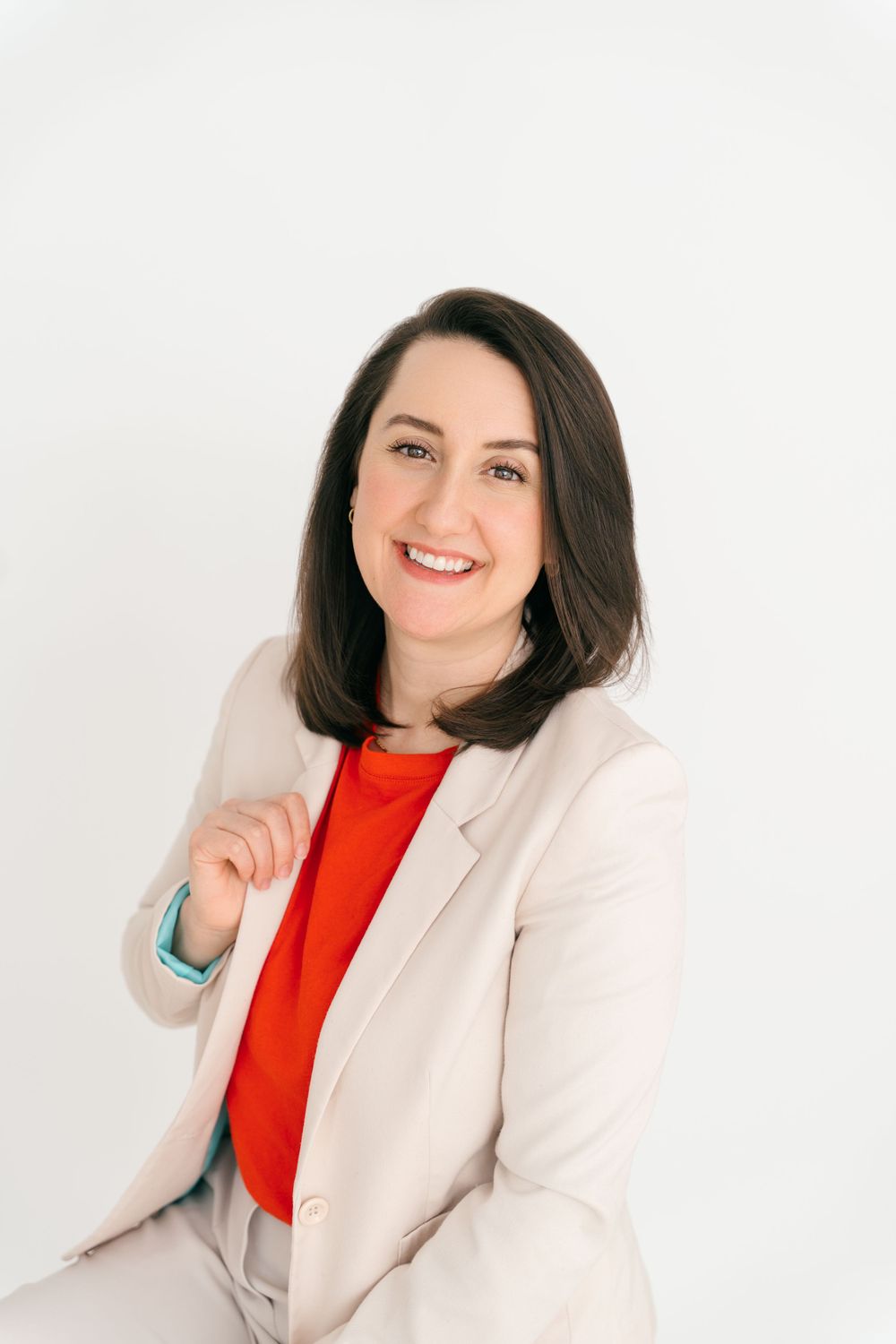 Professional portrait with dark-haired woman in cream blazer and red top smiling against white background.