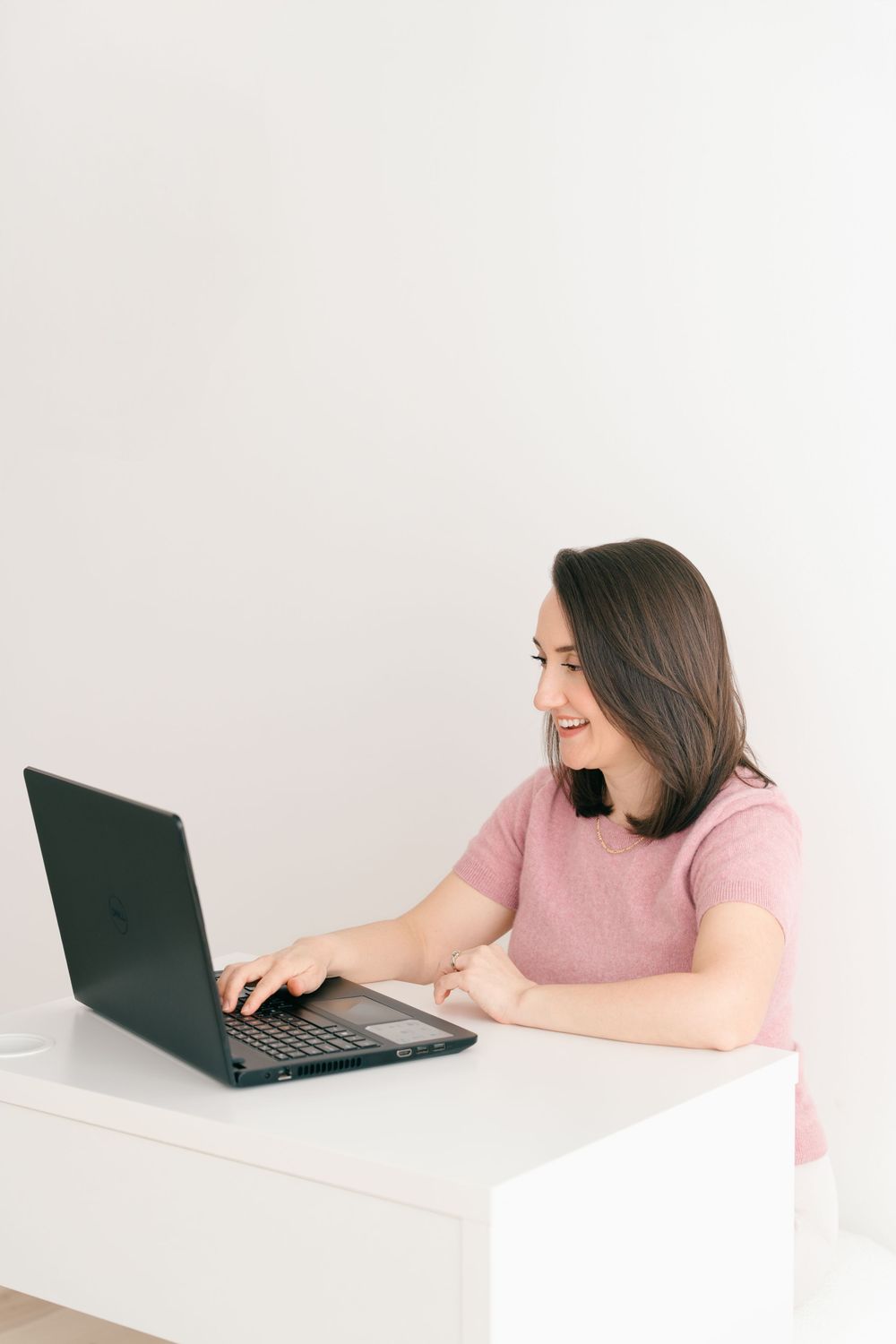 Side view of someone in a pink shirt smiling while working on a laptop at a white desk against light background.