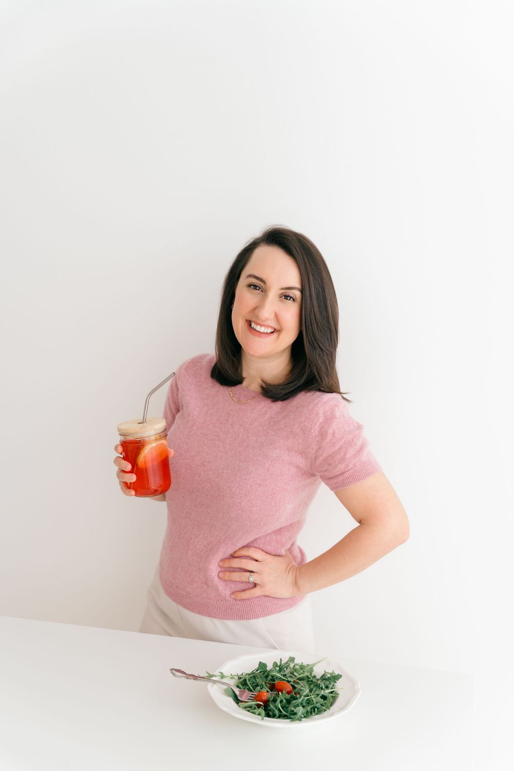 Someone in a pink sweater poses with a bright red cocktail next to a fresh salad on a white table against neutral backdrop.