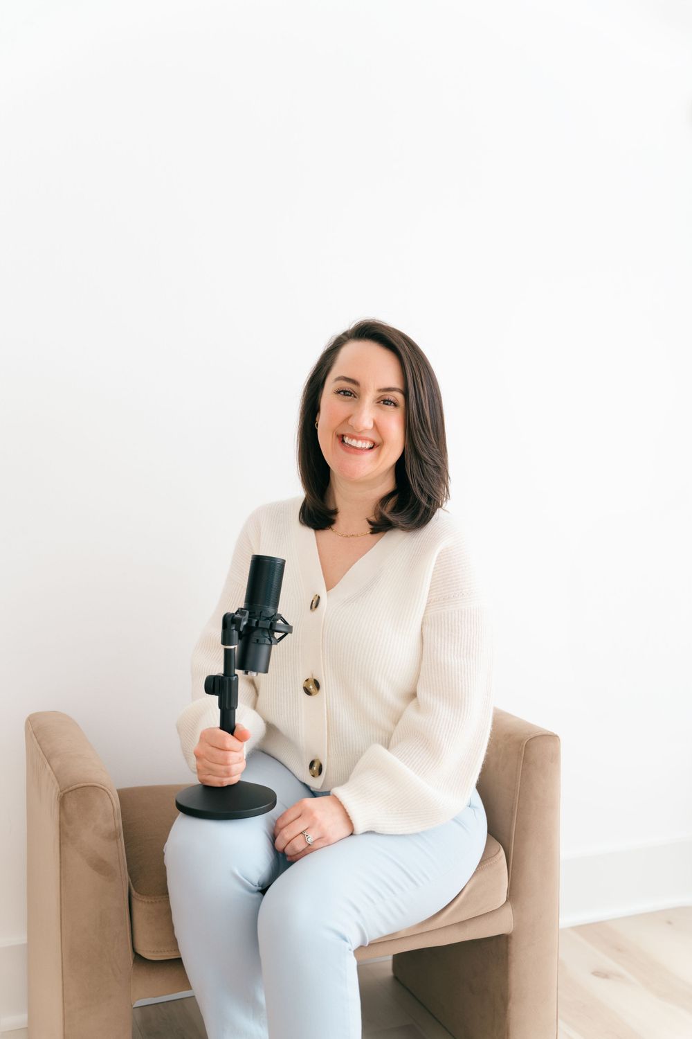 A person in a cream cardigan and light blue pants sits on a beige chair holding a podcast microphone while smiling.