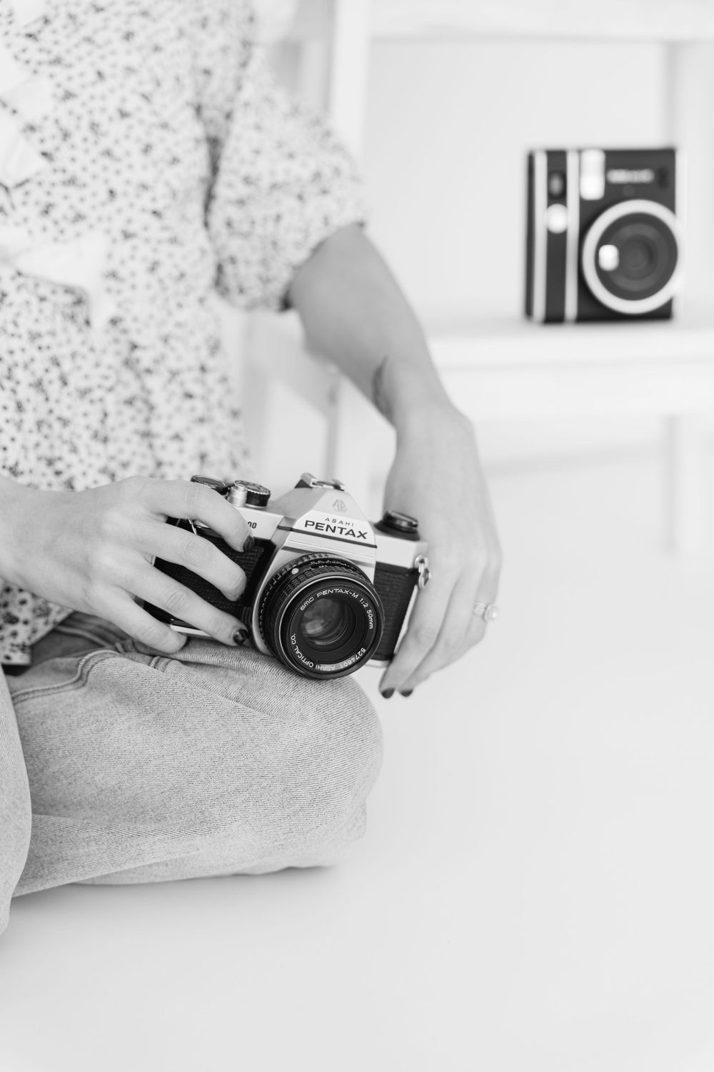 A person holds a vintage Pentax film camera while wearing a polka dot shirt, with another camera visible in background.