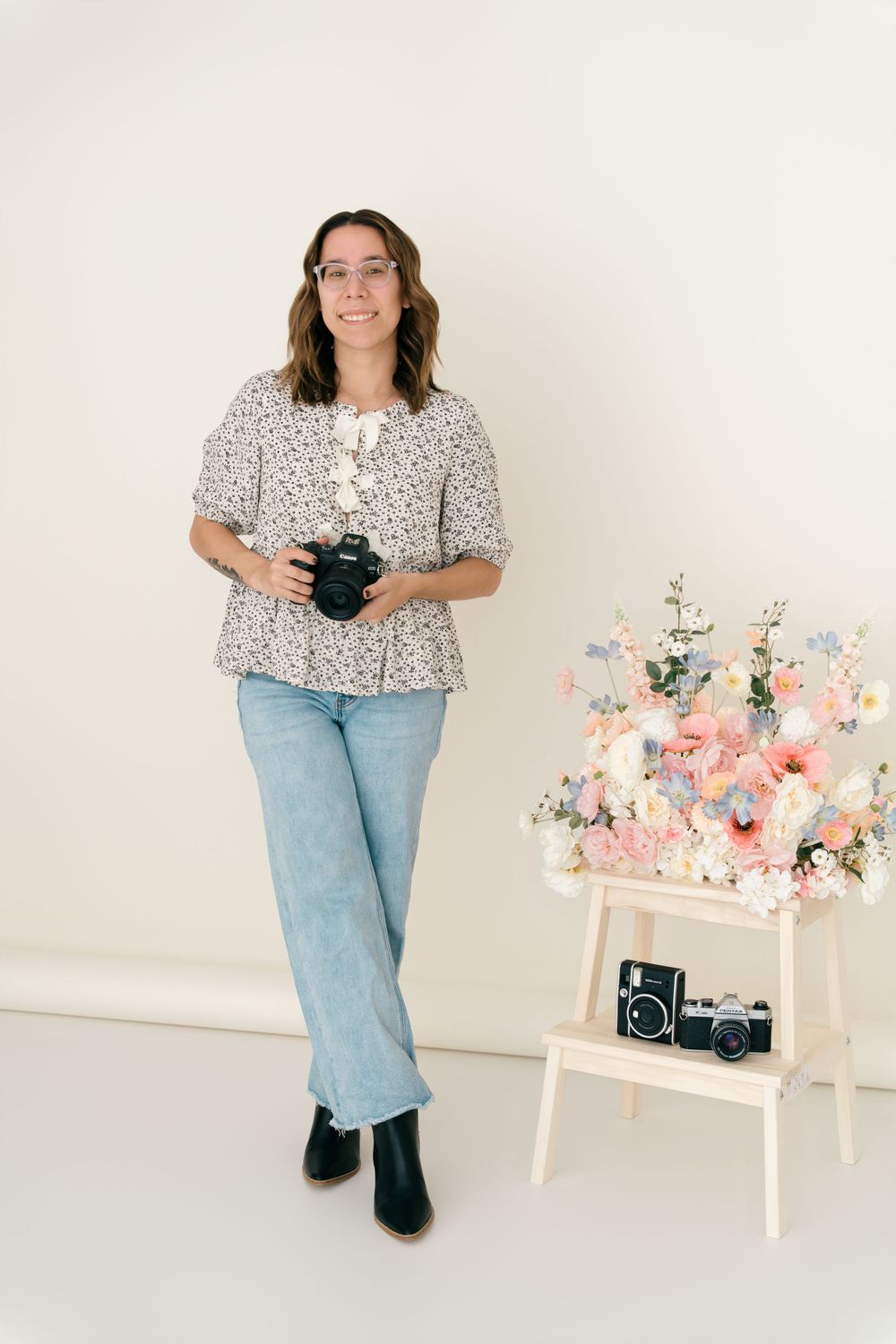 Photographer in floral blouse and jeans holds camera near decorative pink floral arrangement on white ladder shelf.