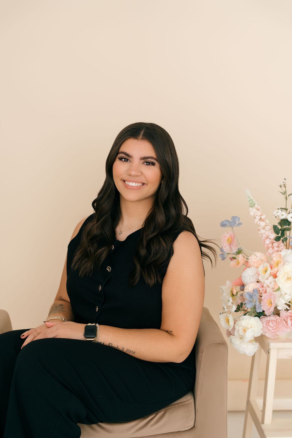A person wearing a black sleeveless dress sits on a beige chair with floral arrangements visible in the background.