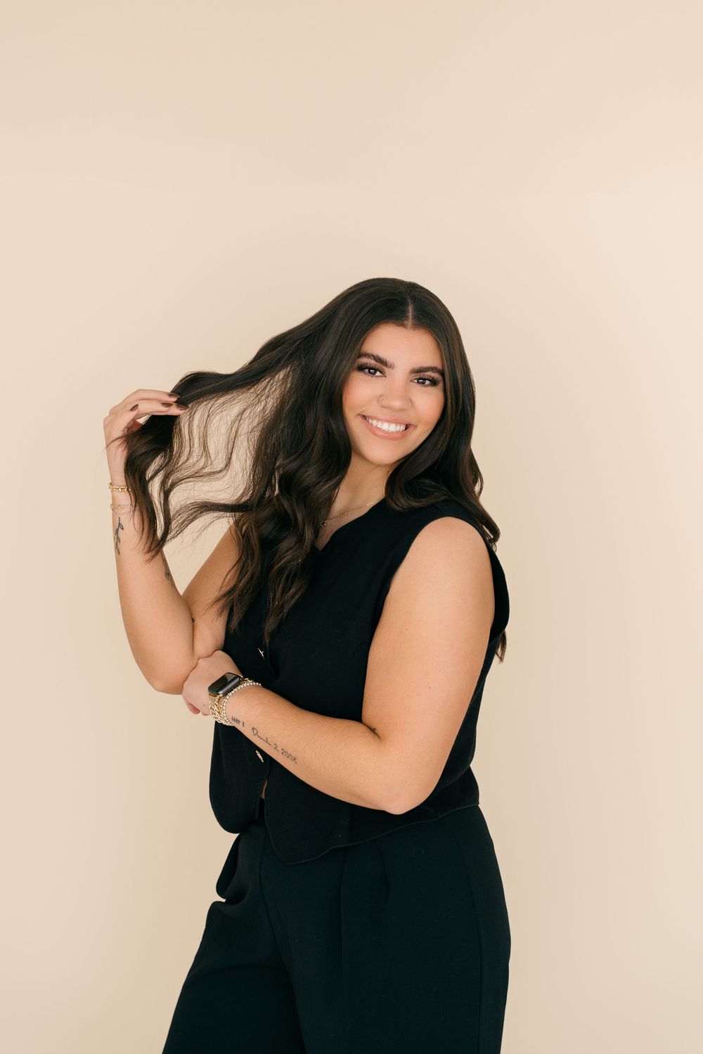 A figure in a black sleeveless outfit poses against a beige background while playfully touching long dark hair.