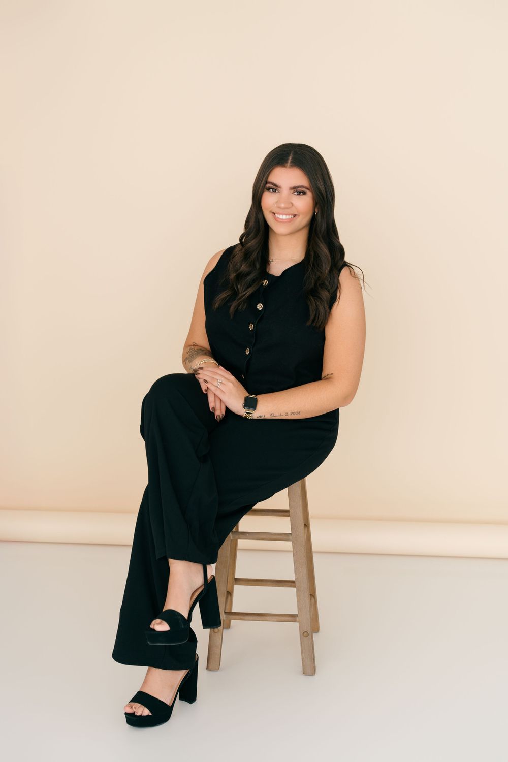 Professional portrait of a woman in a black sleeveless jumpsuit seated on a wooden stool against a light beige background.