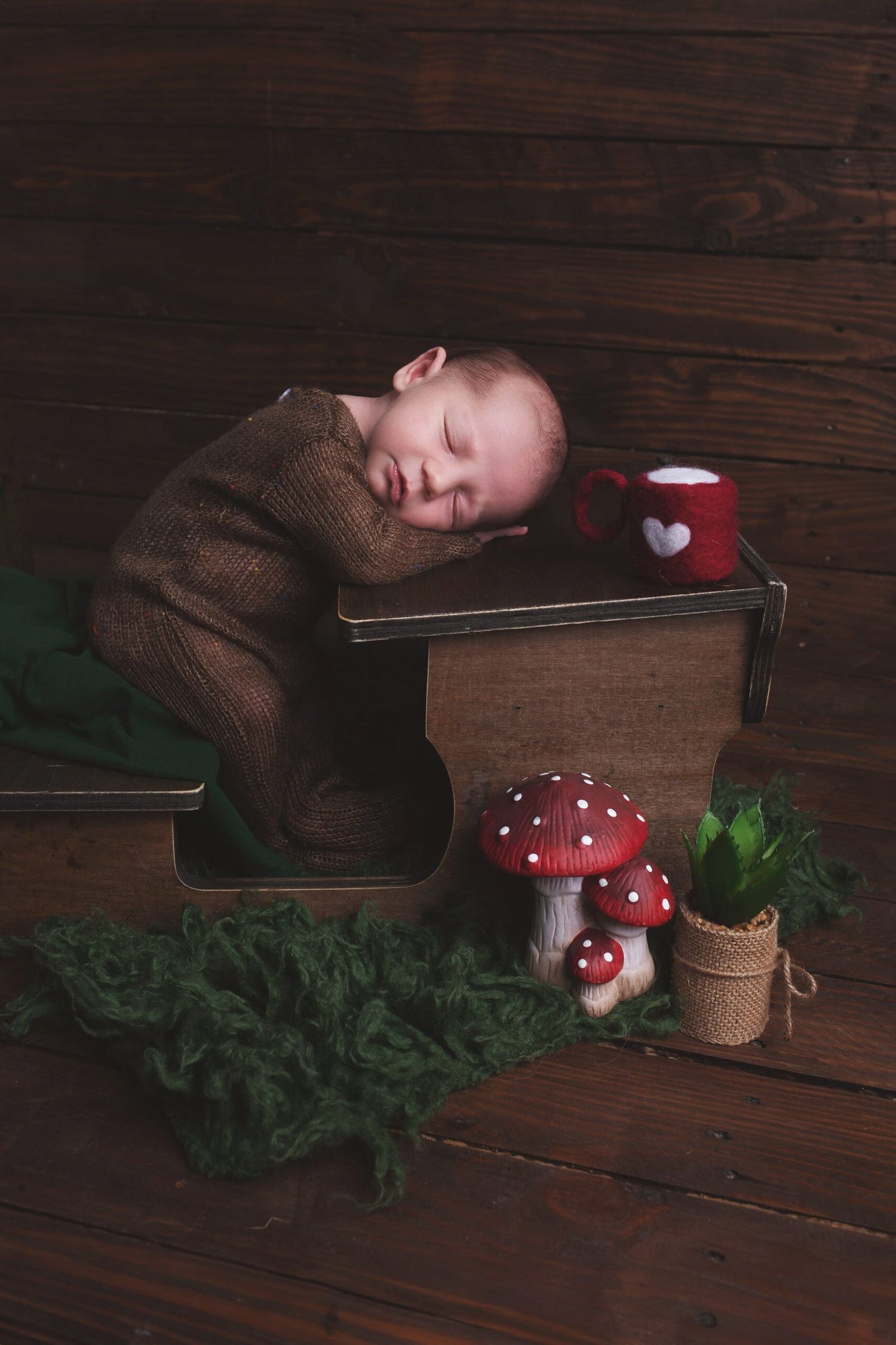 A sleeping baby in brown sweater rests on wooden box with decorative mushroom and holiday decor on rustic wooden floor.