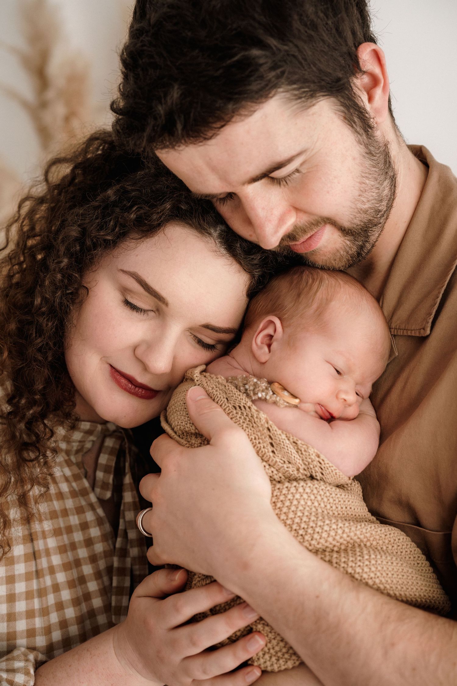 A tender family portrait shows parents in white embracing their newborn baby in a warm, intimate setting.