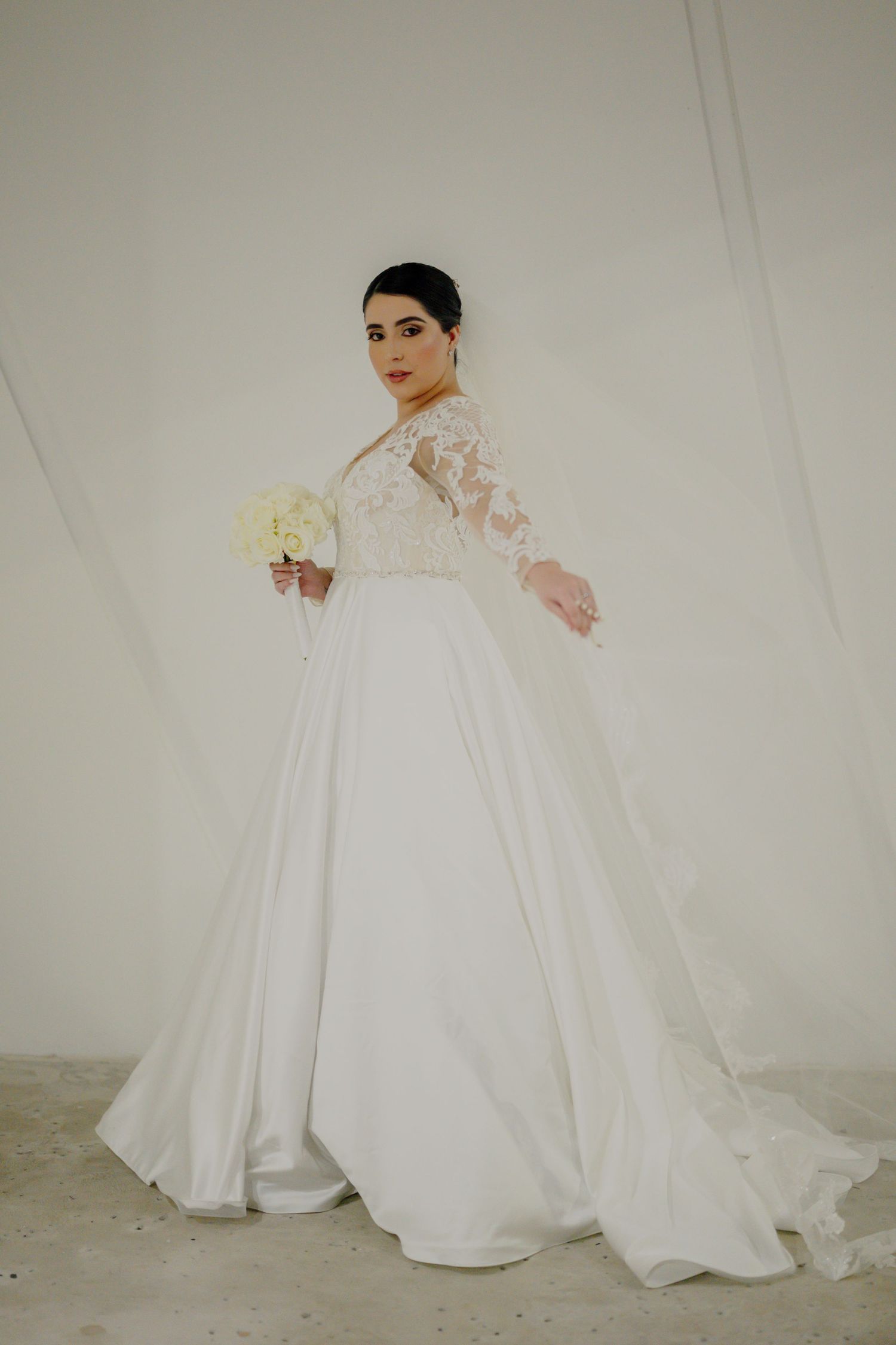 Elegant bridal portrait in flowing white lace and silk ball gown with long sleeves and bouquet against neutral backdrop.