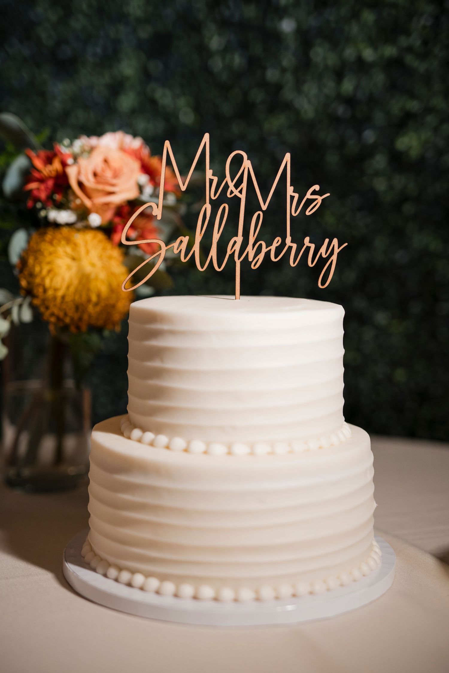 White two-tiered wedding cake with gold 'Mr & Mrs Salldberg' topper and fall floral decor.