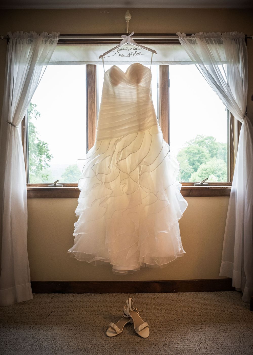An elegant white wedding dress hangs by a window with bridal shoes placed beneath.