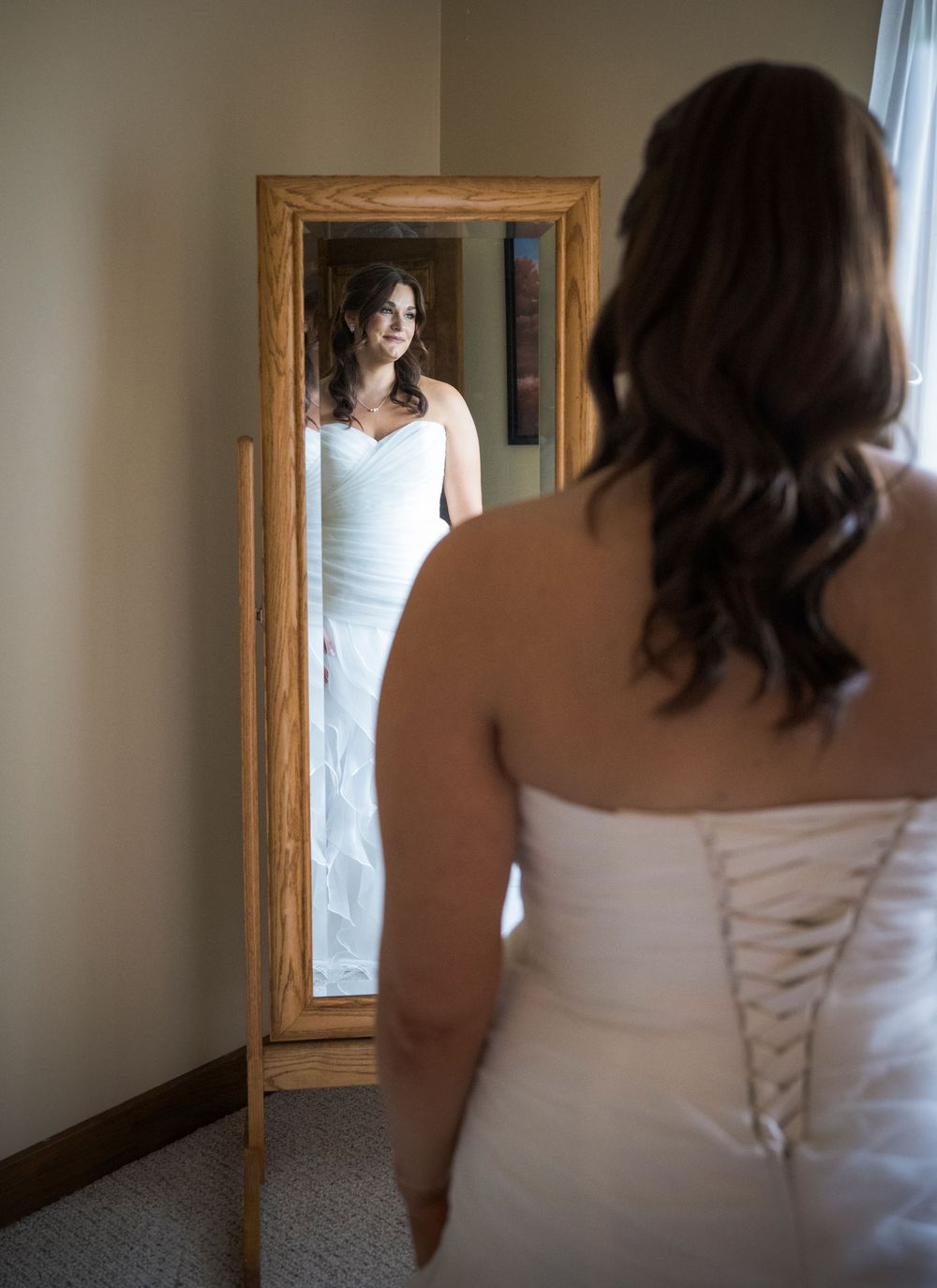 A bride in a white lace-up wedding dress looks at her reflection in an ornate wooden mirror.
