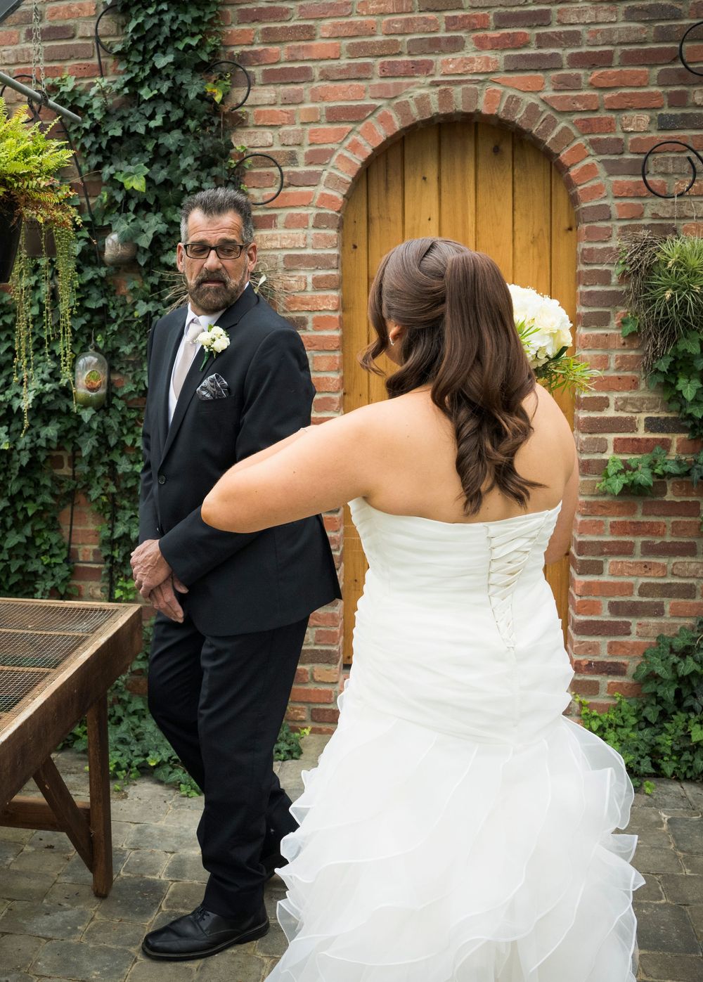 Elegant bride in layered white gown against rustic brick wall with wooden door.