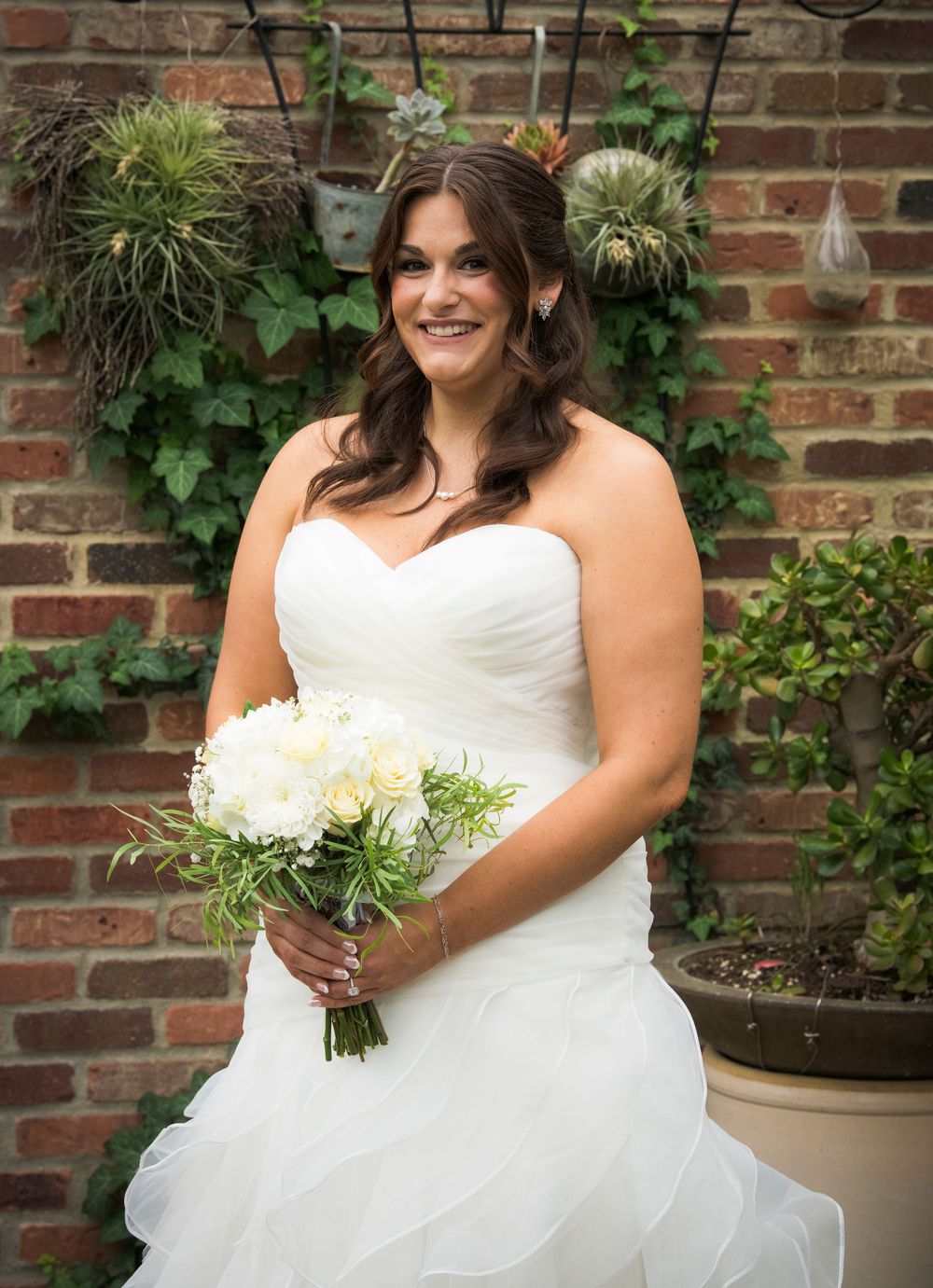 Portrait of bride holding white floral bouquet against brick wall with hanging plants.