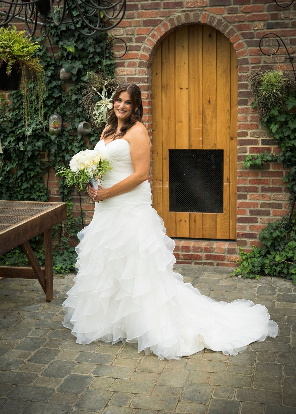 Beautiful white wedding dress with flowing ruffled train photographed against a rustic brick wall and wooden door.