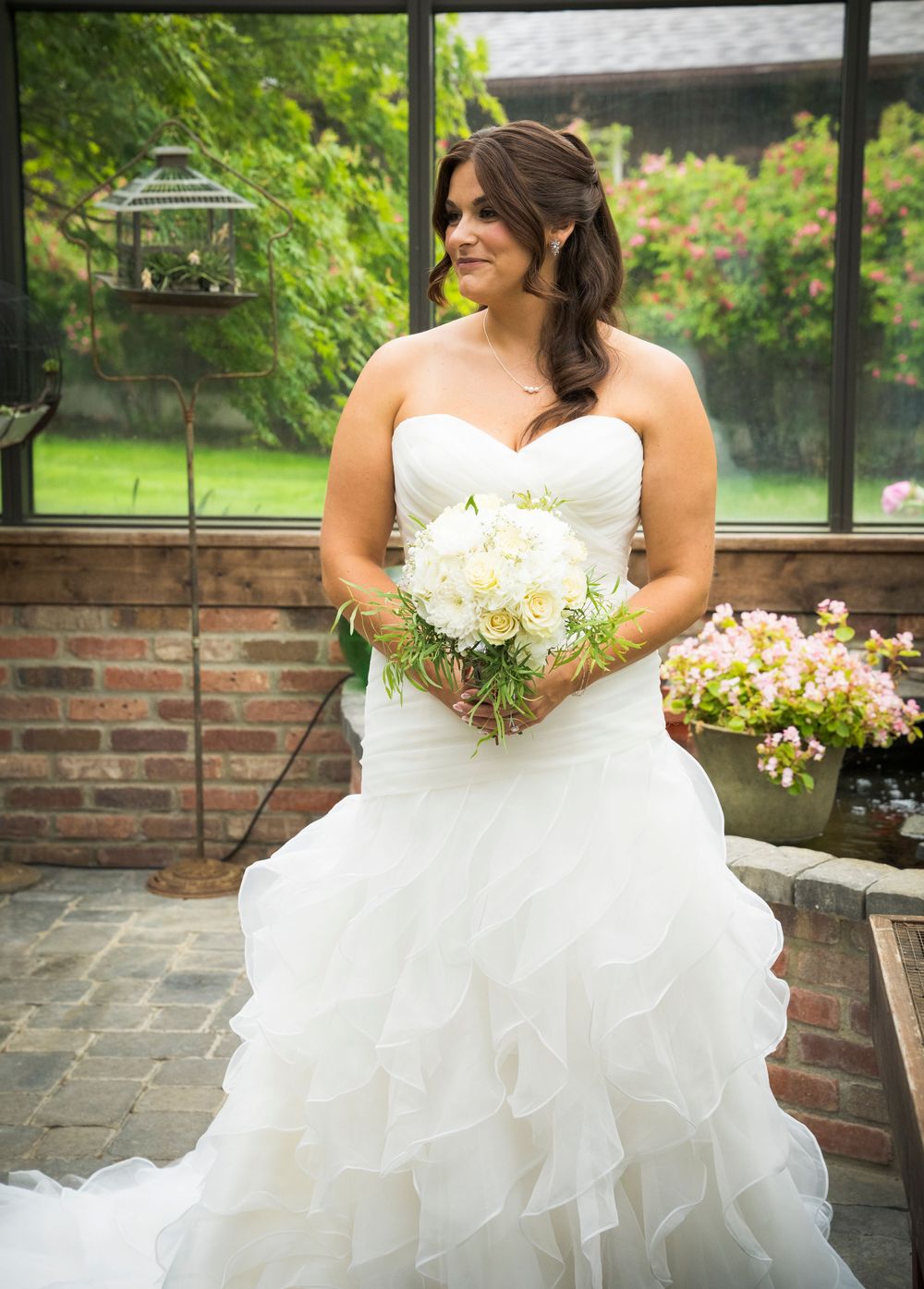 Ruffled white wedding gown and bouquet against garden brick backdrop.