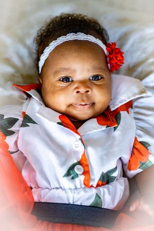 Smiling baby wearing white and red floral outfit with lace headband and flower accessory laying on white bedding.