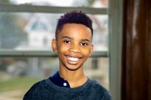 A young person wearing a dark sweater smiles warmly at the camera while standing near a window.
