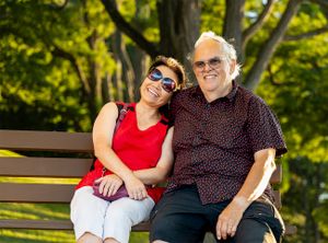 Two people sitting together on a wooden bench in a park with green trees in background on a sunny day.