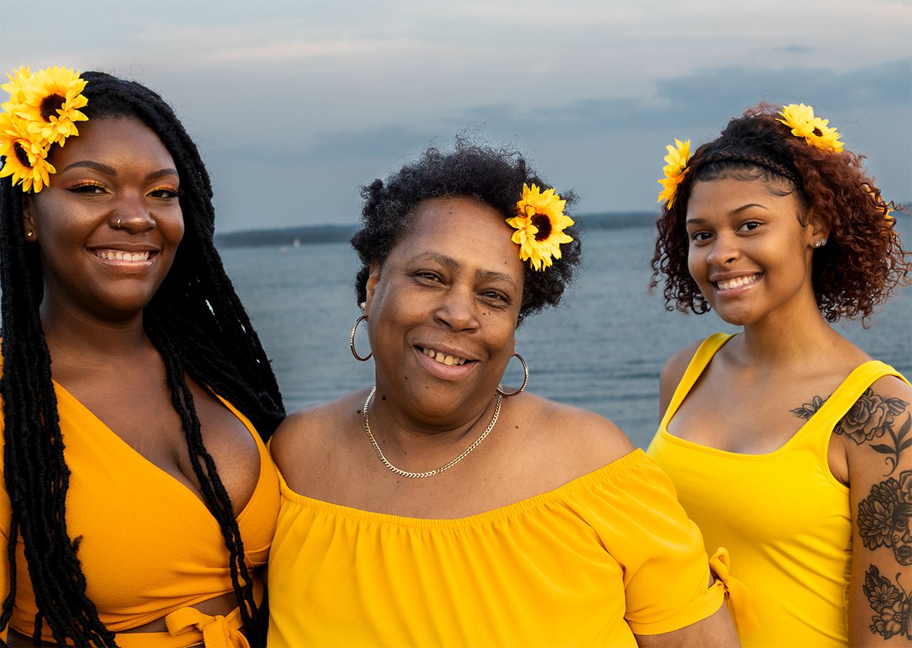 Three people wearing yellow dresses and sunflower hair accessories smile together by the water at sunset.