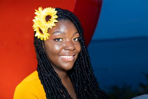 A person with braids wearing a yellow cardigan smiles warmly against a red and blue background with sunflowers in their hair.