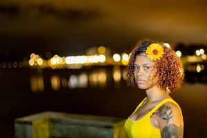 A person in a yellow tank top with curly red hair and a yellow flower accessory poses at night with bokeh lights in background.
