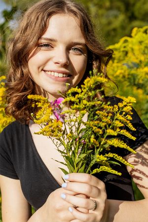 Person in black t-shirt smiling while holding fresh-picked wildflowers including yellow goldenrod on sunny summer day.