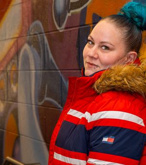 Someone wearing a red, white and blue winter jacket with fur-lined hood and turquoise hair poses against graffiti wall.
