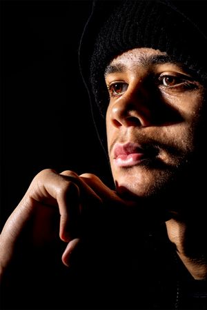 Dramatic portrait in dark lighting shows someone wearing a black beanie with thoughtful expression against black background.