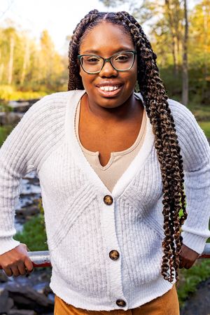 Person wearing a white cardigan sweater and glasses poses outdoors in autumn with long braided hair and bright smile.