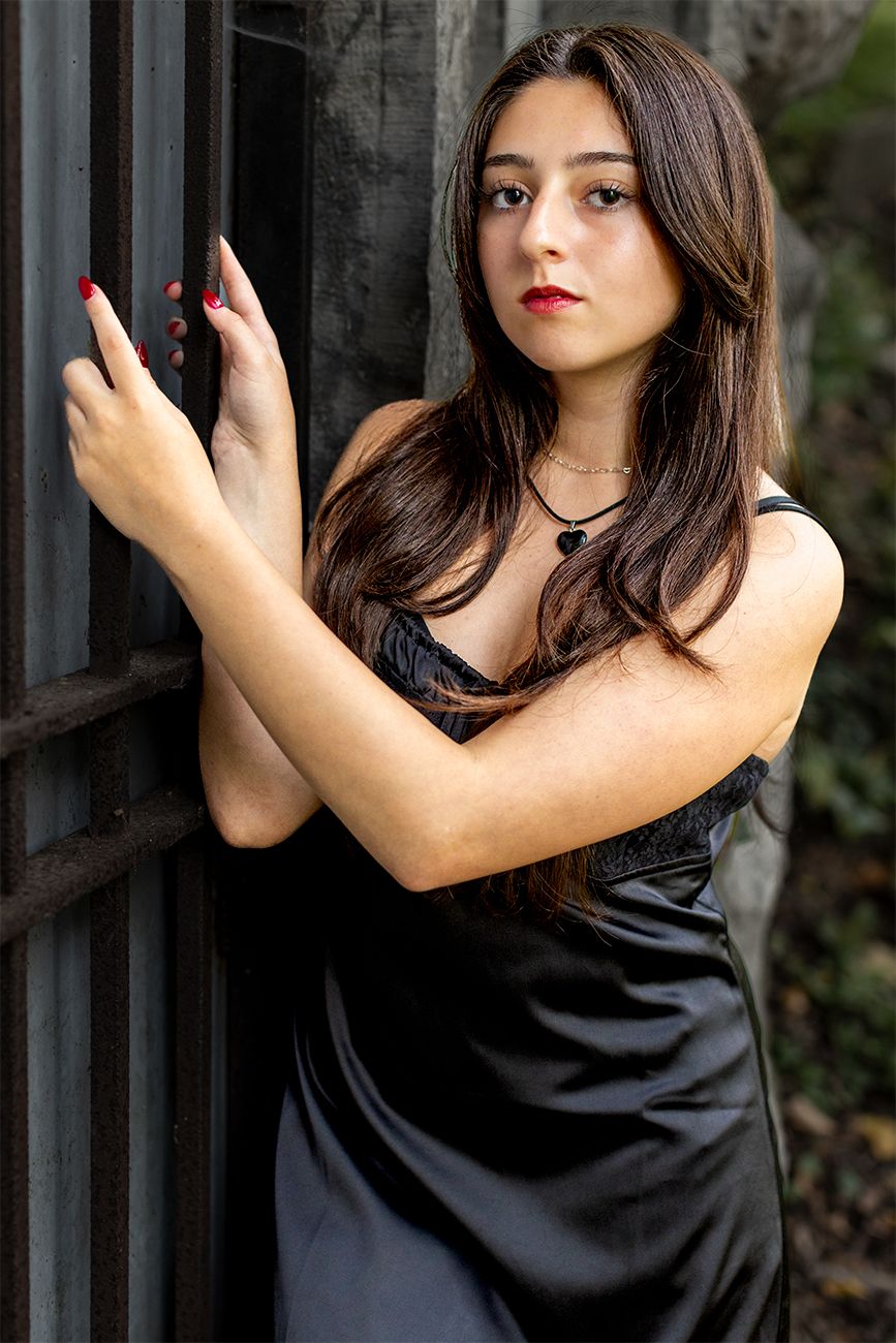 A person in a black satin dress poses elegantly against a dark wall outdoors with red nail polish.