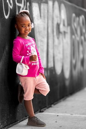 Young child in pink sweater and peach shorts poses against graffiti wall in selective color photography.