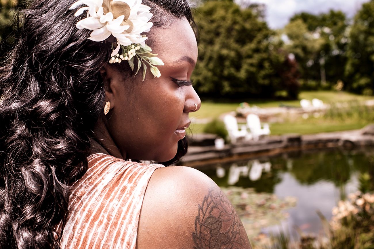 Side profile view of a person with long curled hair wearing a flower hair accessory and sleeveless top near a pond.